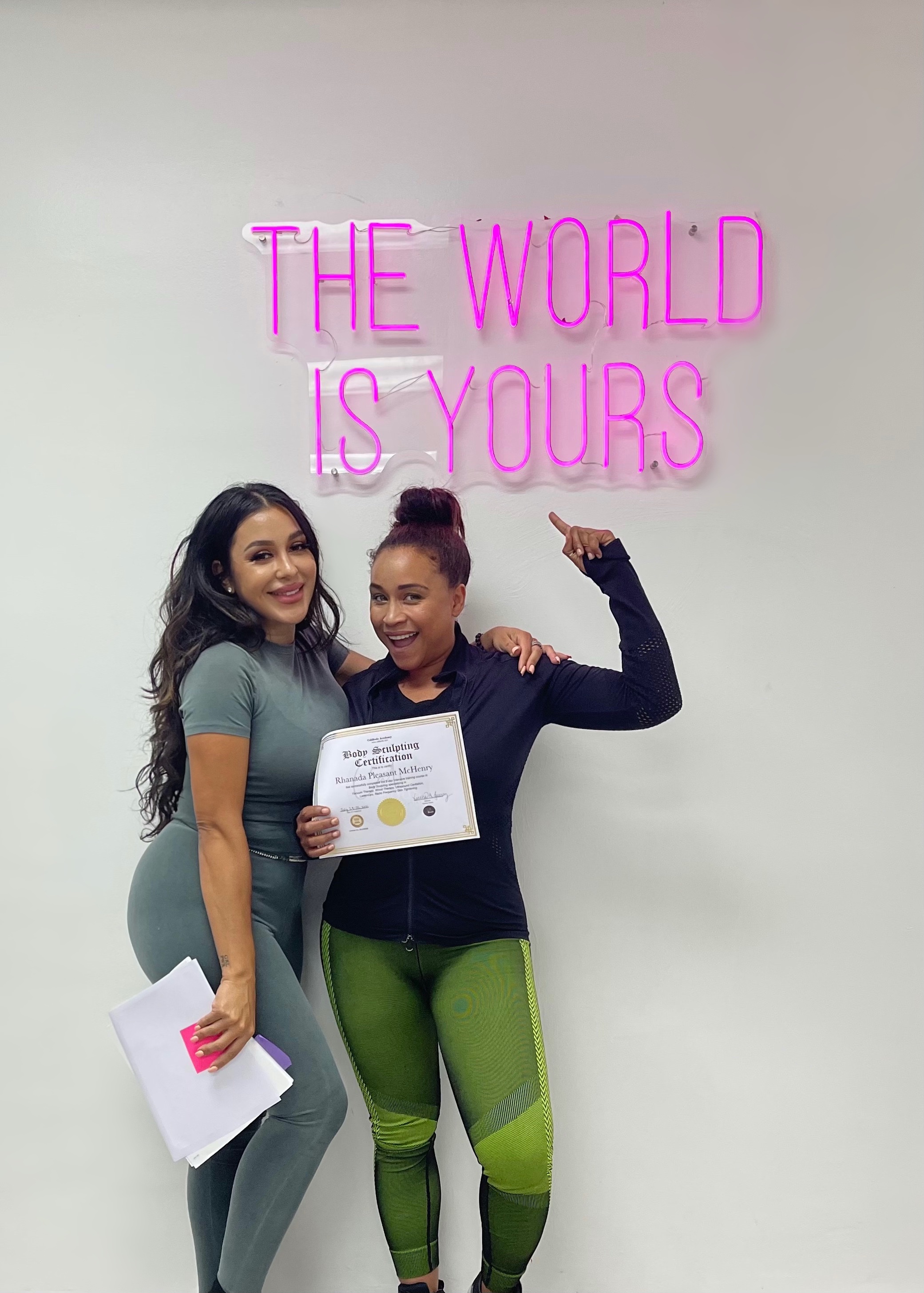 CaliBods academy graduate holding her certificate beside an instructor in front of a neon sign reading The World Is Yours